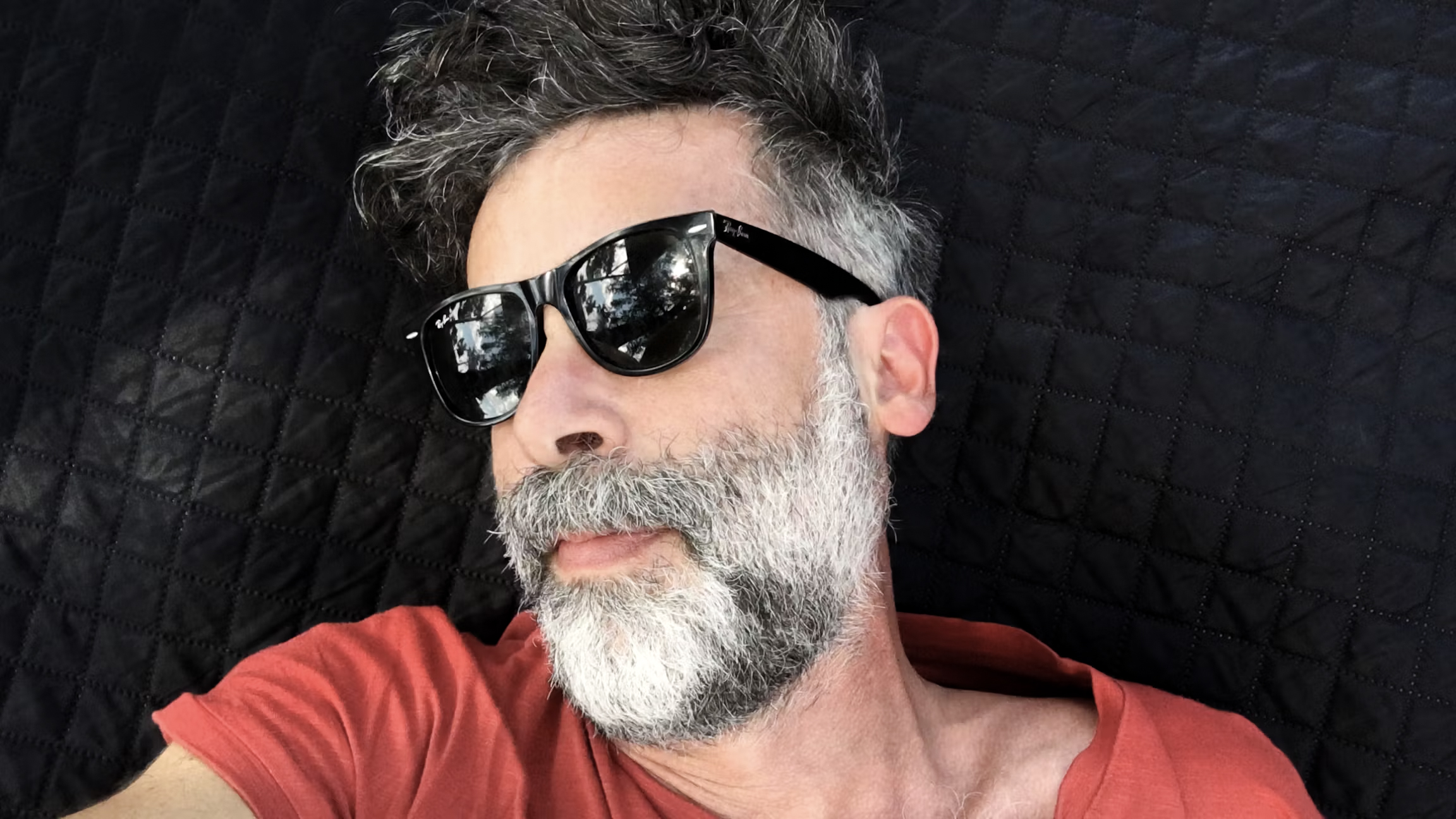 Selbstaufnahme eines Mannes mit Sonnebrille und Vollbart, der auf einer Couch sitzt. 