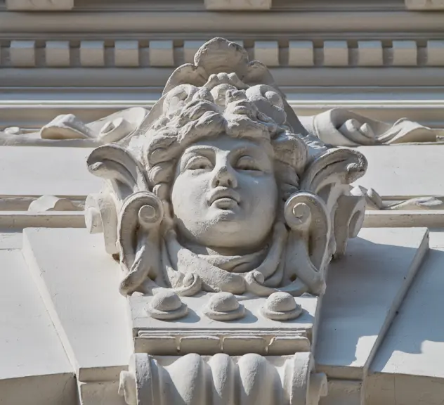 Zu sehen ist ein kunstvolles Element an einer Hausfassade in Form eines Frauenkopfes aus weißem Stein.