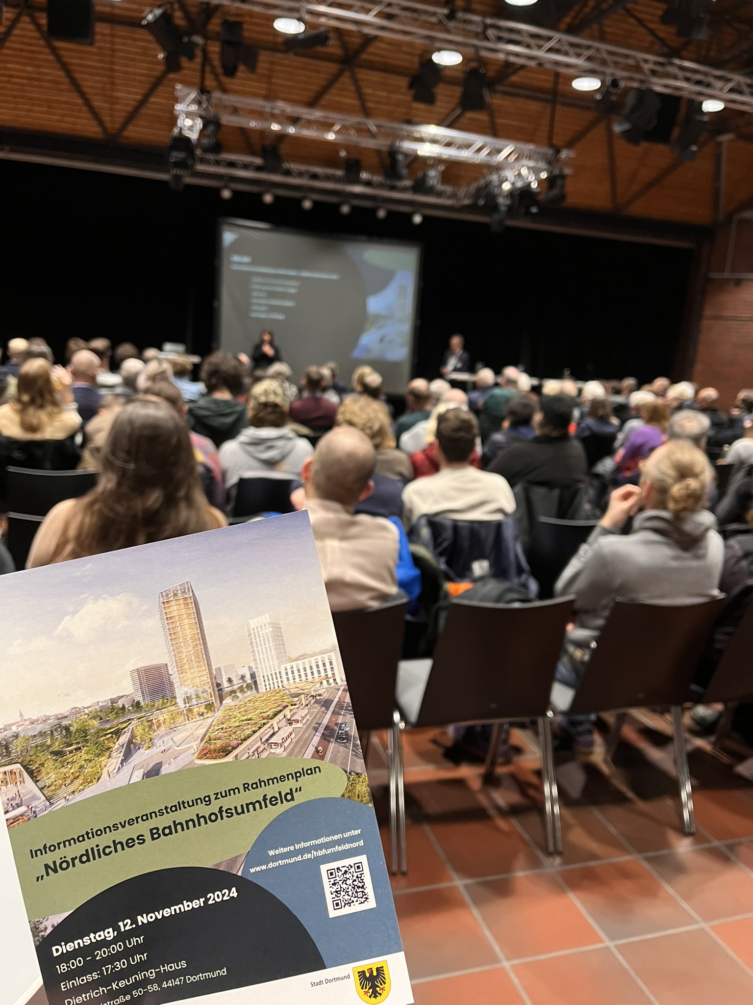 Im Vordergrund der Flyer der Infoveranstaltung, im Hintergrund das Publikum und die Bühne