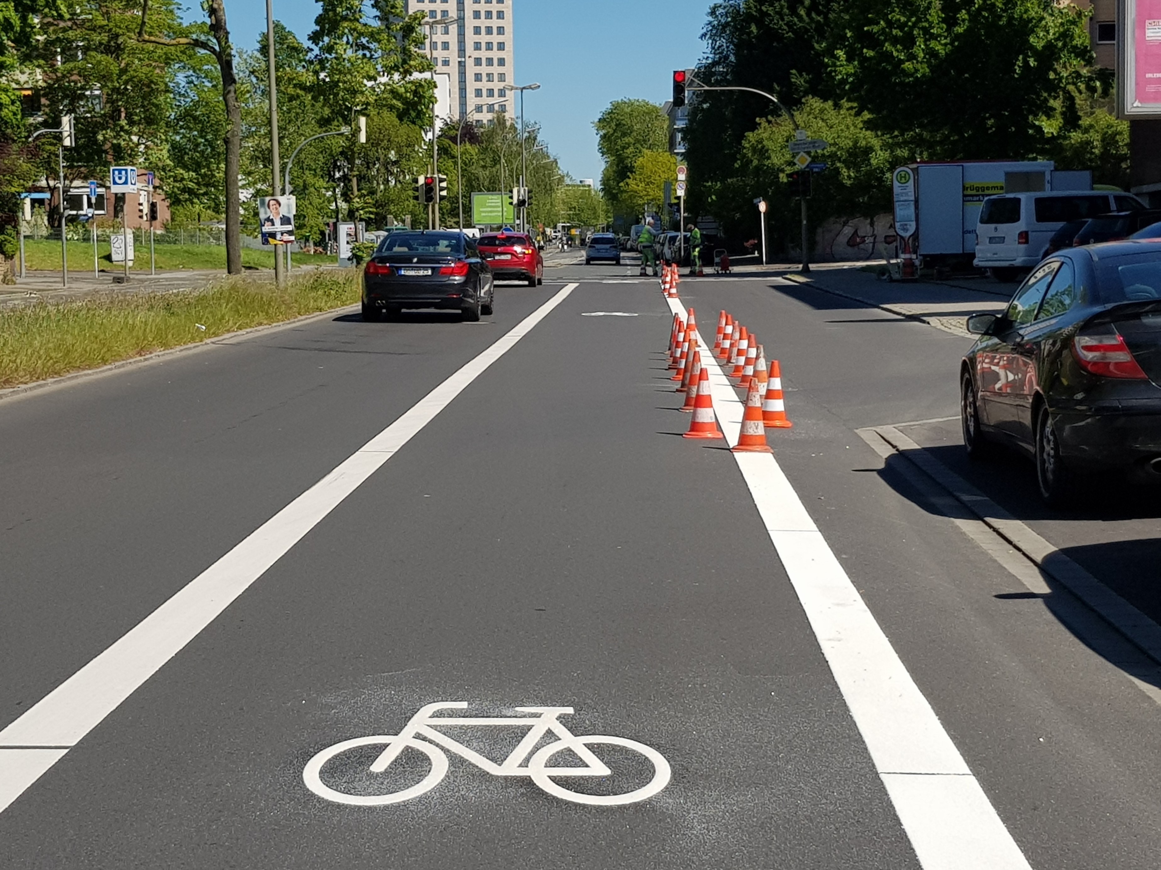 Neumarkierung eines Radfahrstreifens. Im Vordergrund ist ein weißes Fahrradpiktogramm zu sehen. Flankiert wird es von zwei weißen Linien . Links Parken Fahrzeuge. Rechts fahren Fahrzeuge. In der Mitte ist der neue Radfahrstreifen. 