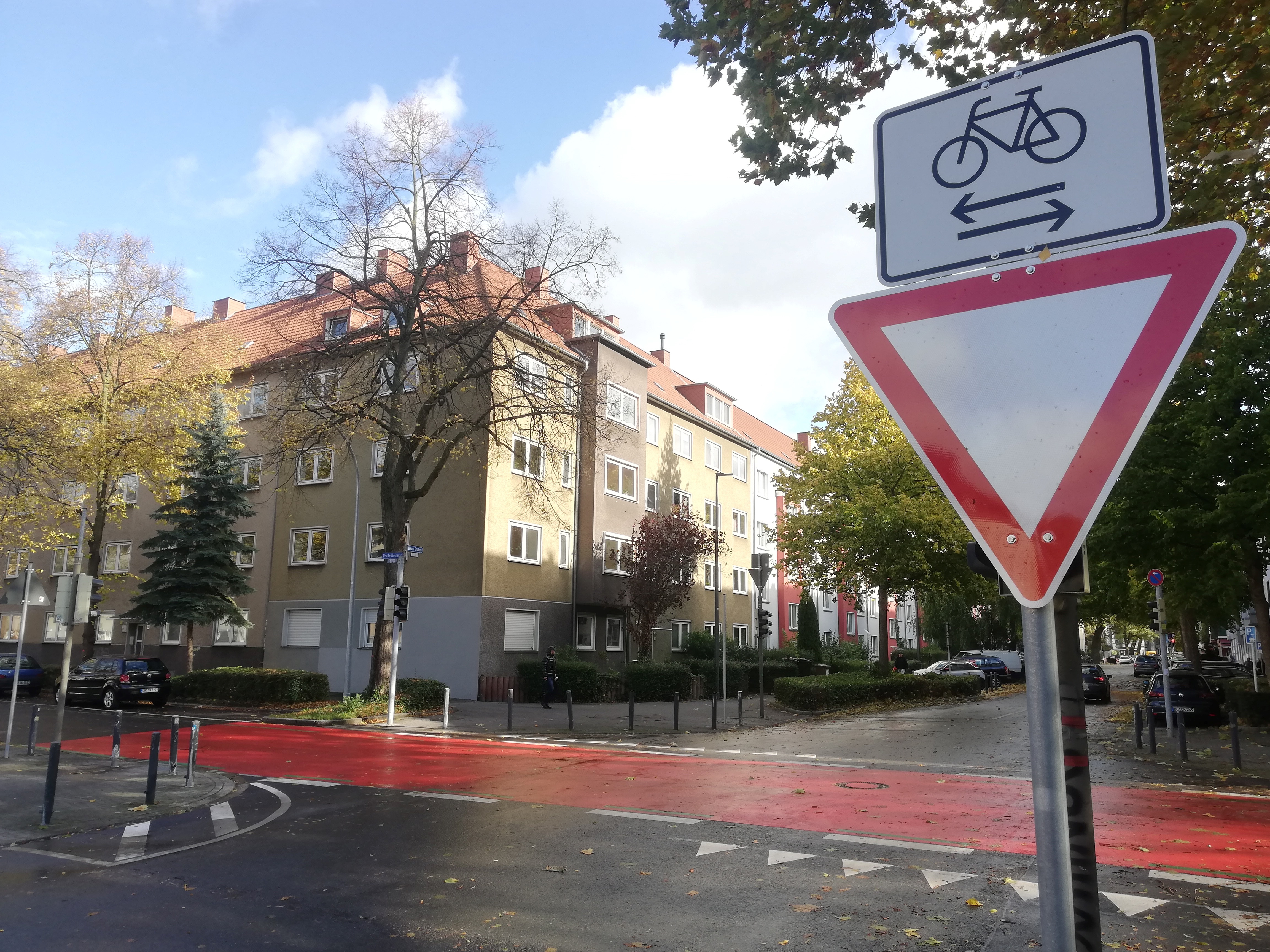 Straßenszene an einer Kreuzung in einem Wohngebiet mit mehrstöckigen Wohnhäusern im Hintergrund. Im Vordergrund sind zwei Verkehrsschilder zu sehen: ein weißes Schild mit einem Fahrradsymbol und zwei entgegengesetzten Pfeilen, das auf Radverkehr in beide Richtungen hinweist, sowie ein darunter angebrachtes „Vorfahrt gewähren“-Schild. Die Fahrbahn ist im Kreuzungsbereich rot markiert, was auf eine Fahrradstraße oder einen Fahrradüberweg hinweist. Bäume säumen die Straßenränder, und es ist herbstlich, mit Laub auf dem Boden und teils kahlen Bäumen.
