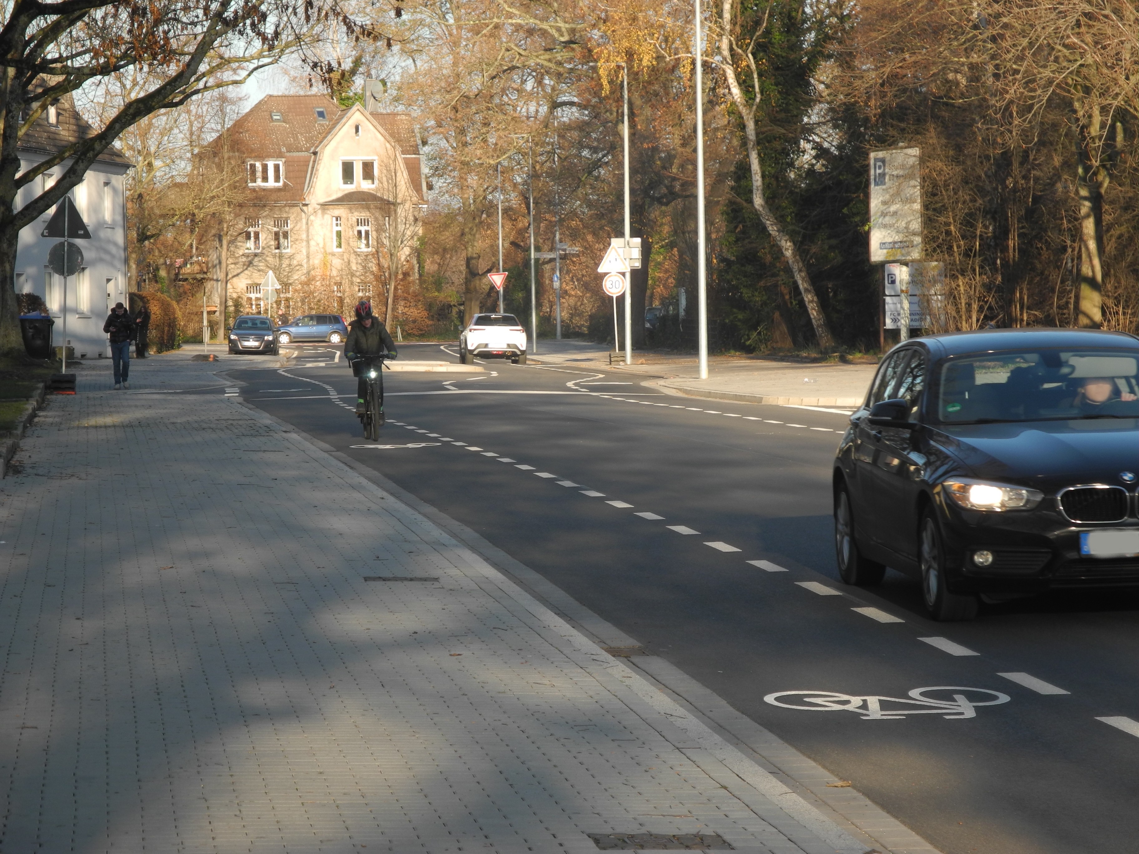 Fahrradfahrende Person auf einem Fahrradschutzstreifen. Im Vordergrund fährt ein Auto. 