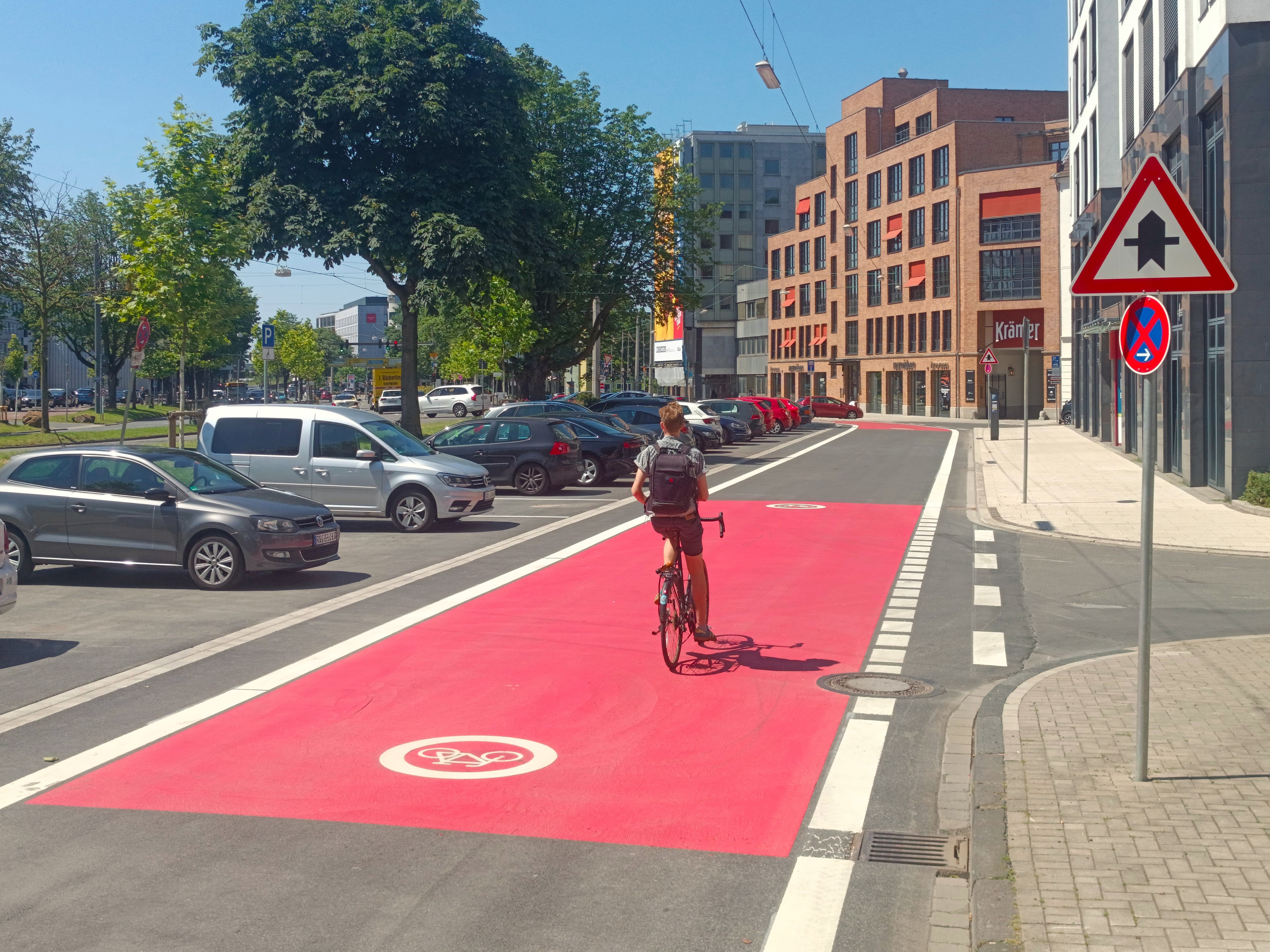 Städtische Straßenszene mit einer deutlich markierten Fahrradstraße, die im Kreuzungsbereich rotmarkiert ist. Mittig fährt ein Radfahrer in sportlicher Kleidung. Links der Straße befinden sich parkende Autos, rechts moderne Büro- und Geschäftsgebäude. Verkehrsschilder weisen auf die Vorfahrt an der Kreuzung sowie ein absolutes Halteverbot hin. Es herrscht sonniges Wetter mit blauem Himmel.