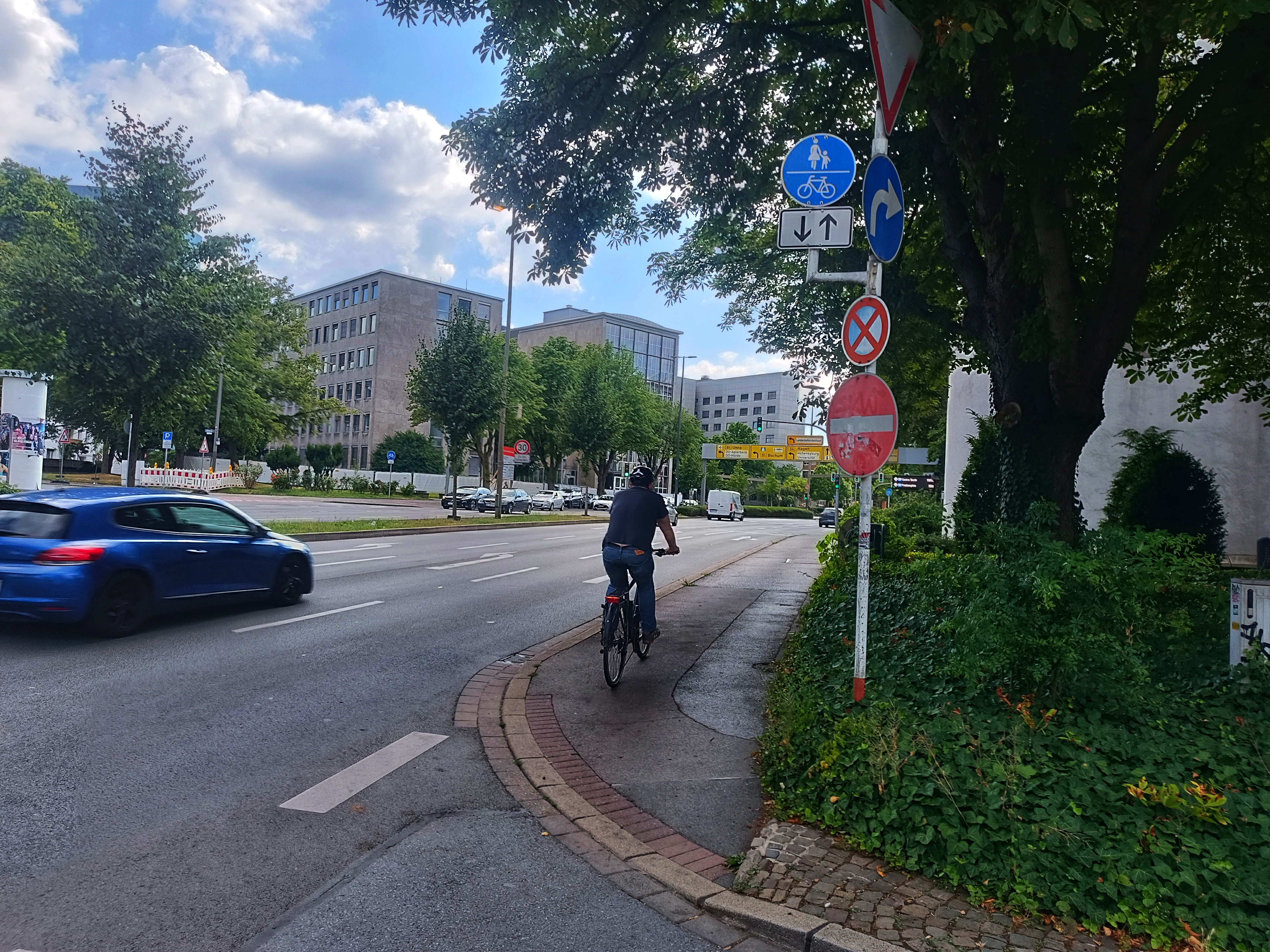 Ein Radfahrender fährt neben einer Straße auf dem Radweg. Rechts im Bild steht ein Mast mit Schildern. 