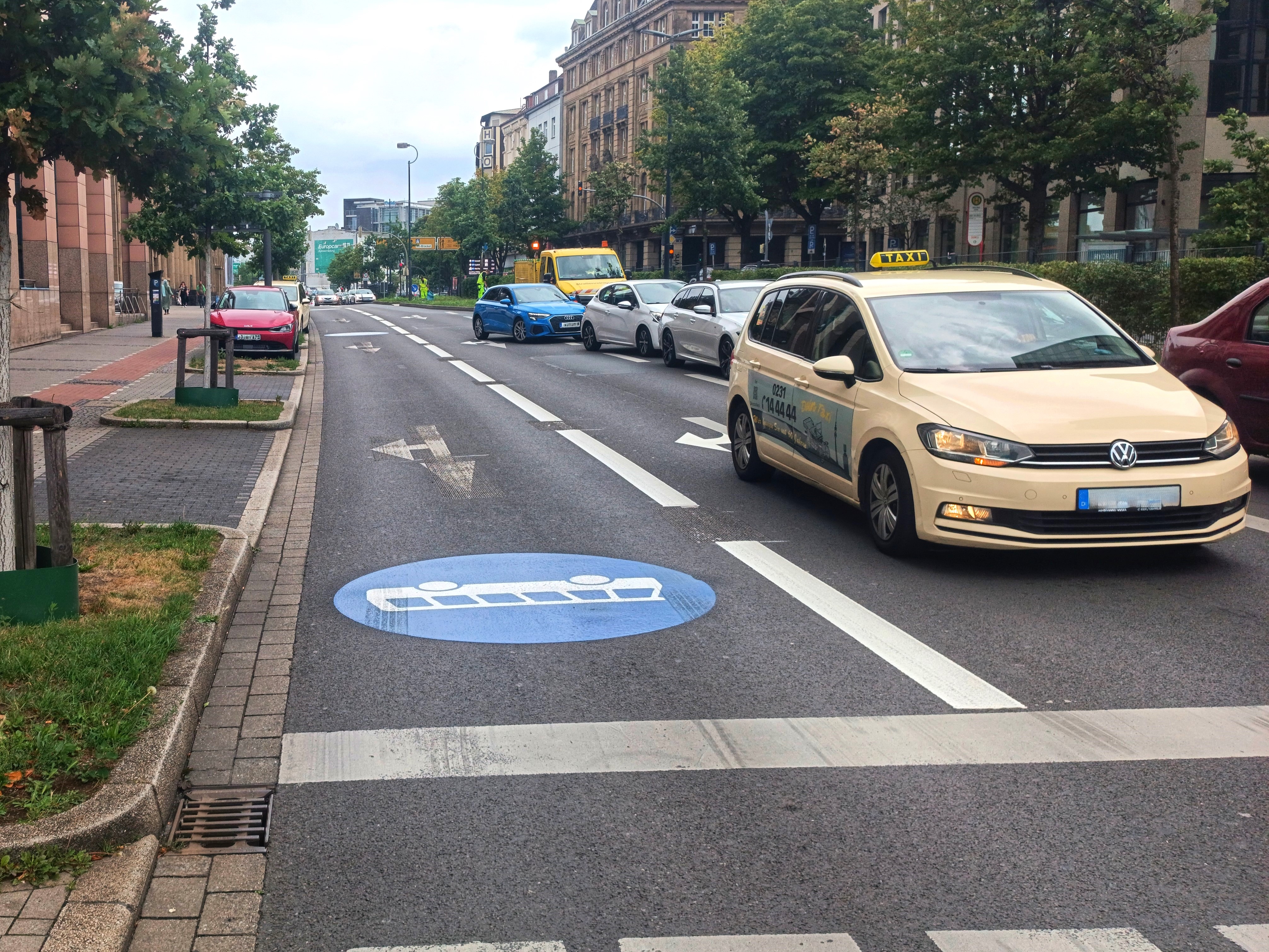 Straßenszene in einer Stadt mit mehrspuriger Fahrbahn. Auf der rechten Spur ist ein blaues Piktogramm für eine Busspur auf den Asphalt gemalt. Mehrere Fahrzeuge, darunter ein beiges Taxi im Vordergrund, fahren auf der anderen Spur. Links am Straßenrand stehen geparkte Autos, daneben verläuft ein Geh- und Radweg mit Bäumen. Im Hintergrund sind weitere Autos, ein gelber Lieferwagen und mehrstöckige Gebäude zu sehen.