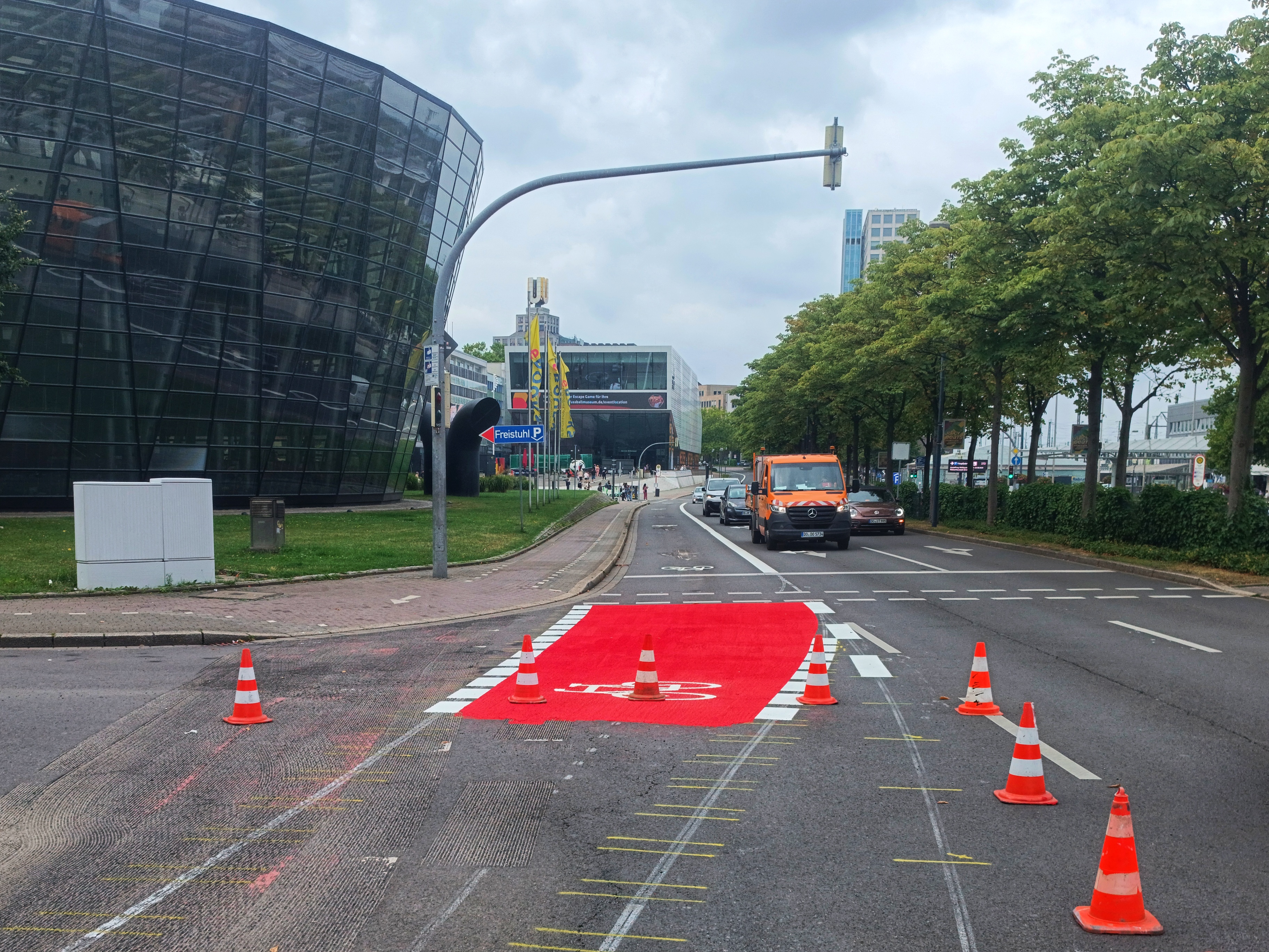 Stadtstraße mit frisch markiertem, rotem Radfahrstreifen in der Mitte der Fahrbahn, umgeben von mehreren orange-weißen Verkehrskegeln. Links im Bild steht ein modernes, schräg verglastes Gebäude, rechts eine Baumreihe entlang der Straße. Im Hintergrund fahren ein orangefarbener Wartungswagen und weitere Autos stadteinwärts. Fußgänger sind in der Ferne vor einem modernen Gebäudekomplex mit Glasfassade und Werbebannern zu sehen.