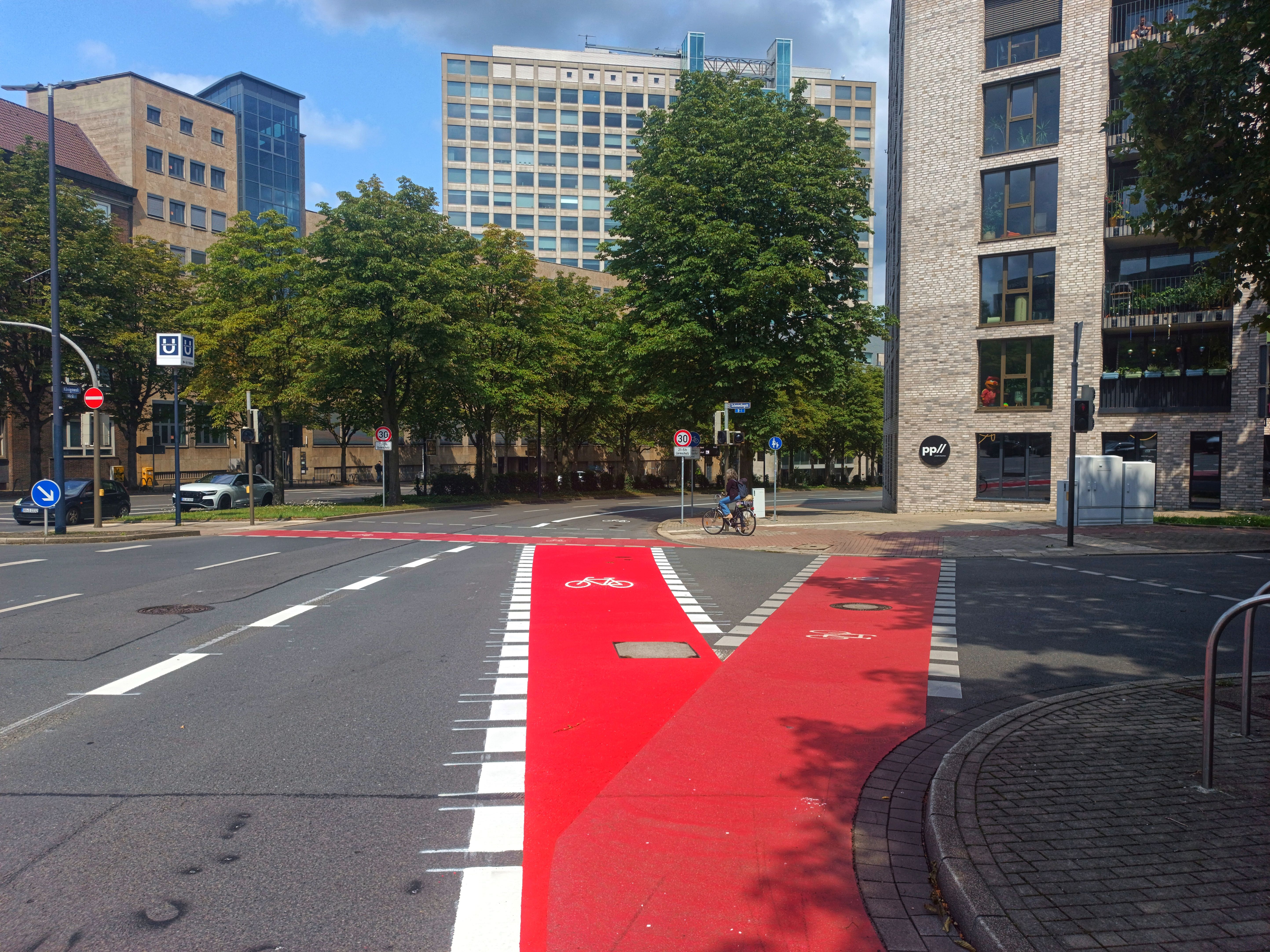 Städtische Kreuzung mit mehreren Fahrspuren und Radwegmarkierungen. Im Vordergrund sind zwei rote Fahrradfurten zu sehen, links daneben verläuft eine Fahrspur. Im Hintergrund sind Bürogebäude und Bäume zu sehen. In der Mitte des Bildes befindet sich eine Radfahrerin, die an der Kreuzung auf grün wartet. 