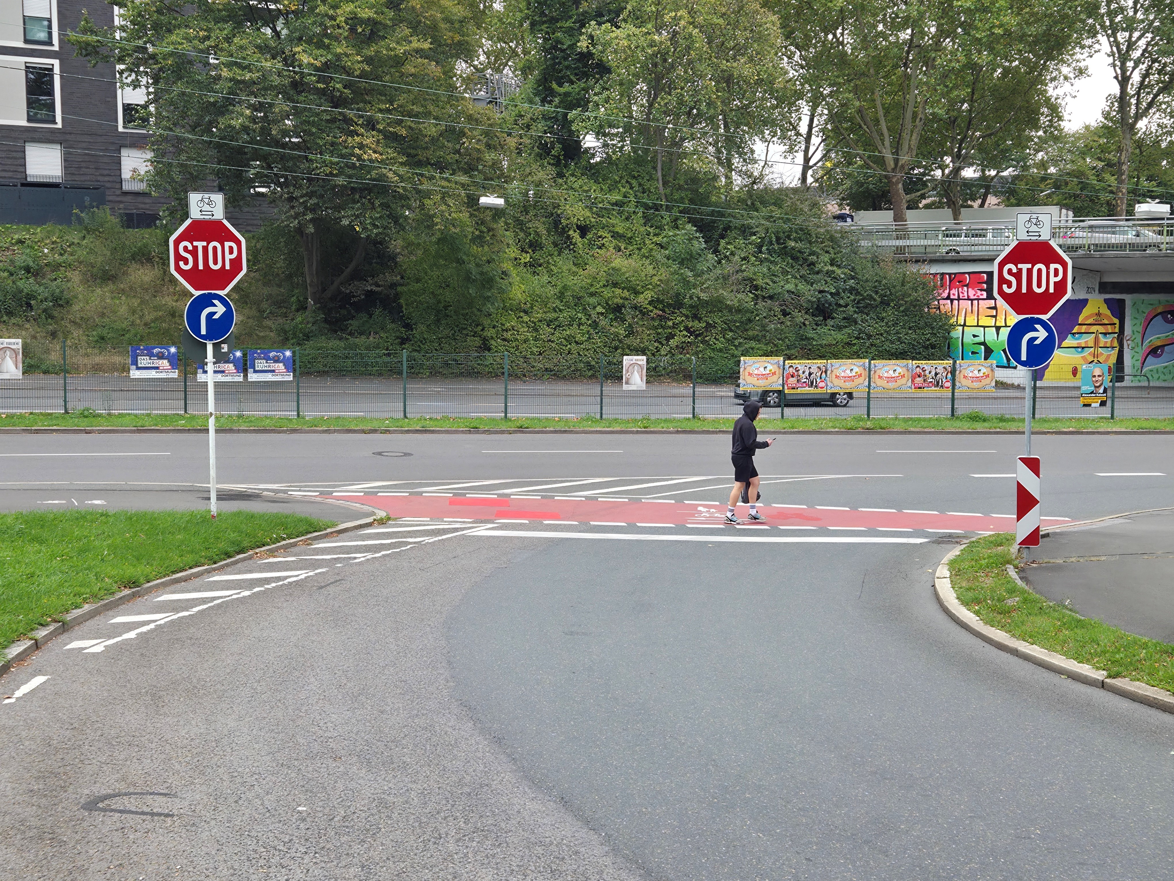 Eine untergeordnete Straße mündet in einer querende Straße ein. Links und rechts stehen Stopschilder mit Zuzusatzzeichen, dass mit Radverkehr in beiden Richtungen zu rechnen ist. Die Furt der querenden Geh- und Radweges ist rotmarkiert.