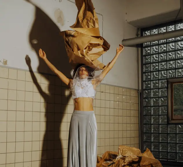 Eine Szene aus dem Stück- Eine Frau steht mit erhobenen Armen in einer alten Halle und hat einen riesigen Hut aus braunem Papier.